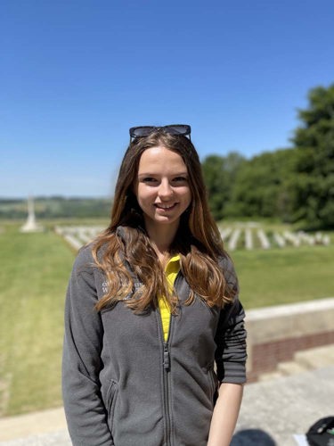 Eleanor poses in a grey CWGF jacket and yellow CWGF polo shirt in front of the graves at the Thiepval Memorial