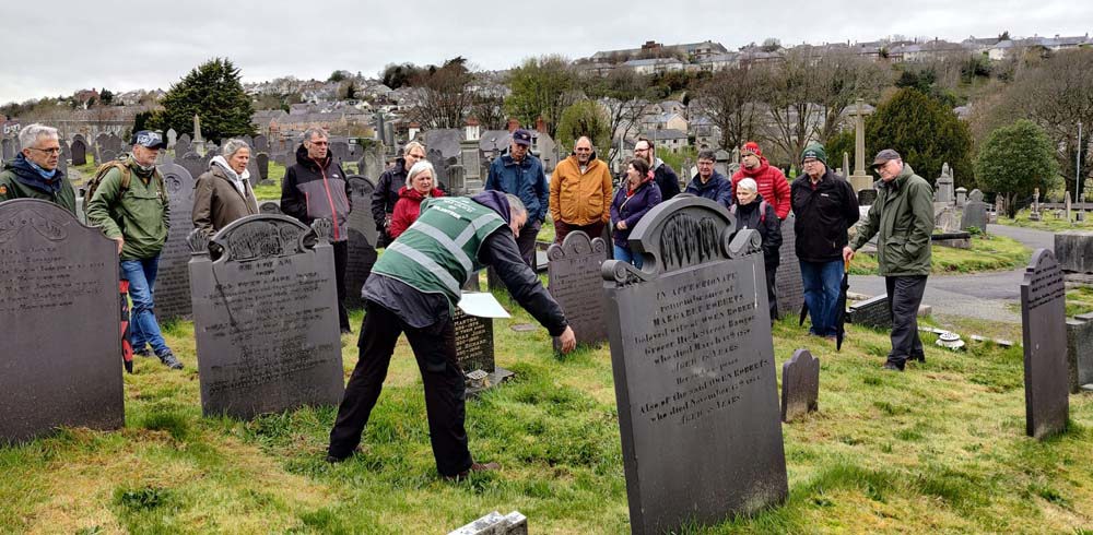 CWGC Volunteer Tour Guide Adrian Hughes, pictured in the green hi vis vest, delivers a talk to a mixed audience of tour goers in a Welsh cemetery. Adrian is pointing to a war grave. Several dark headstones can be seen in the cemetery.