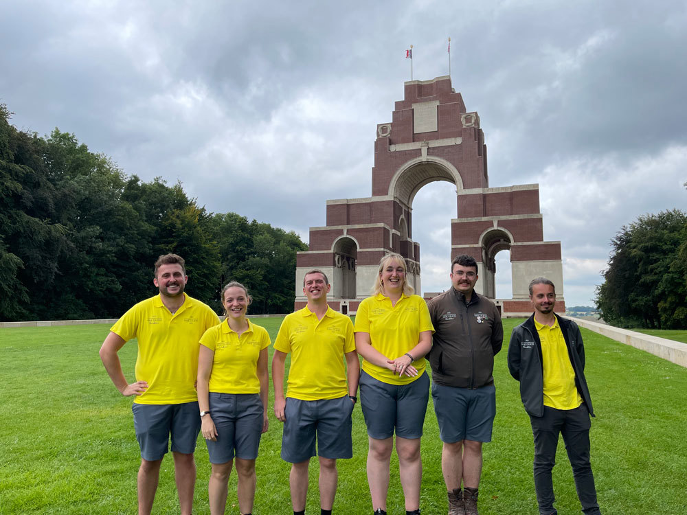 CWGF Guides posing in front of the Thiepval Memorial. The group of young people are all dressed in yellow polo shirts and mostly shorts, with some sporting light jackets.