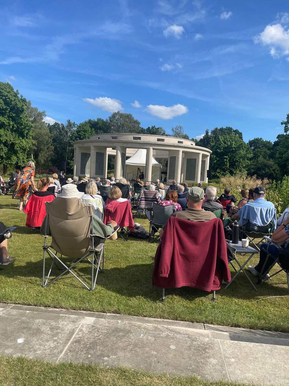 Concertgoers watch a harpist playing on the rotunda of the Brookwood Memorial in Brookwood Military Cemetery. The white stone rotunda is flanked by various species of green tree under a blue sky featuring wispy white clouds.