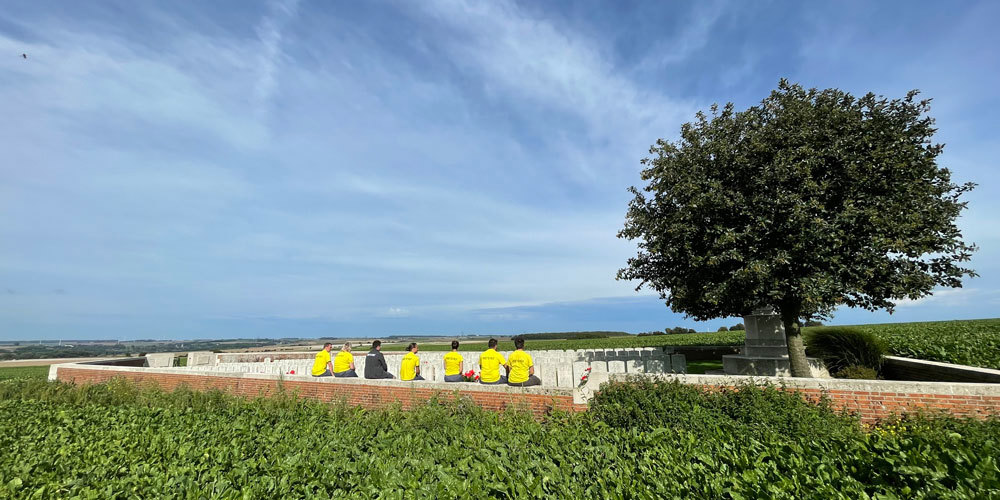 CWGF guides in bright yellow poloshirts sitting on the red brick wall of a CWGC cemetery in France. A large tree is visible to the right.