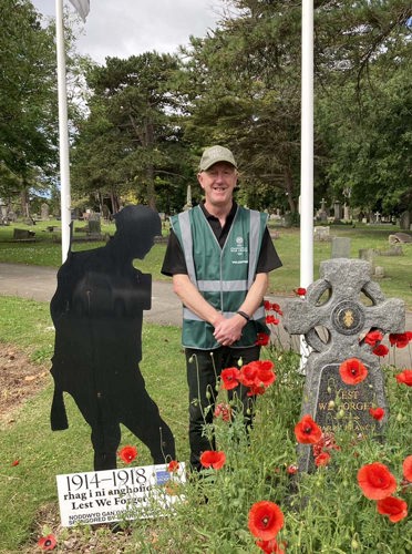 Man in a green hi-vis jacket stands next to a silhouette statue of a WW1-era soldier and a Celtic Cross-shaped headstone. Poppy's have been planted around the headstone.