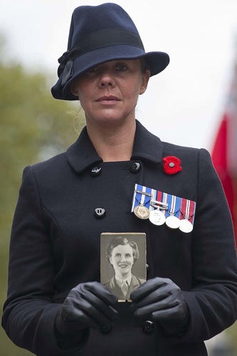 A womean in black coat and dark hat holds up a picture of a ATS casualty. The woman is wearing a poppy and has several RAF medals pinned to her breast.