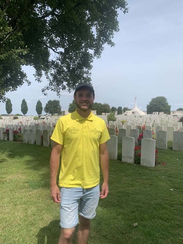 Portrait of Luke Goddard standing in front of headstones at Tyne Cot Cemetery/ He is wearing a yellow polo, grey hat, and light denim jean shorts.