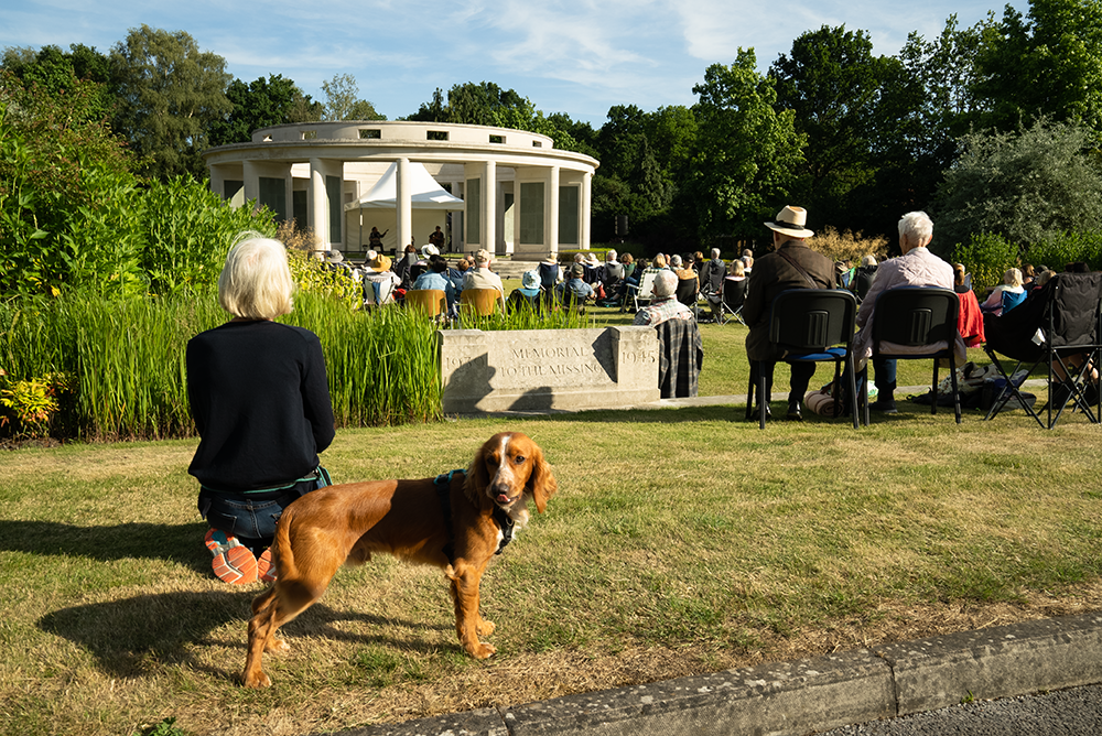 A crowd, with a brown spaniel in the foreground, watch performers play music on the central rotunda of the Brookwood 1939-1945 War Memorial in Brookwood Military Cemetery.