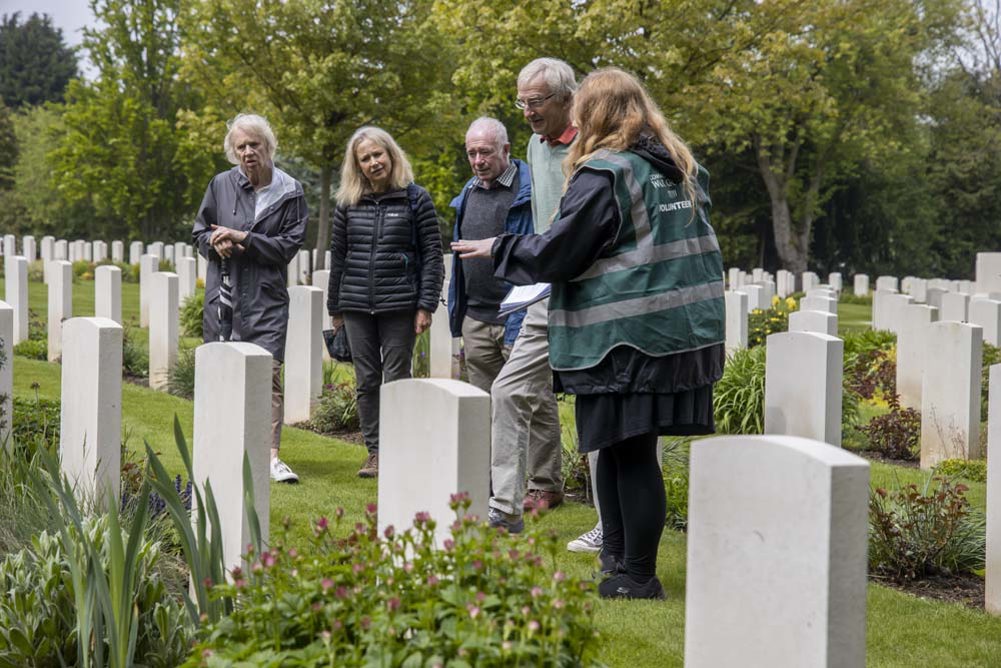 A tour guide with long brown hair, wearing a dark green reflective vest, speaks to a tour group in a CWGF cemetery.