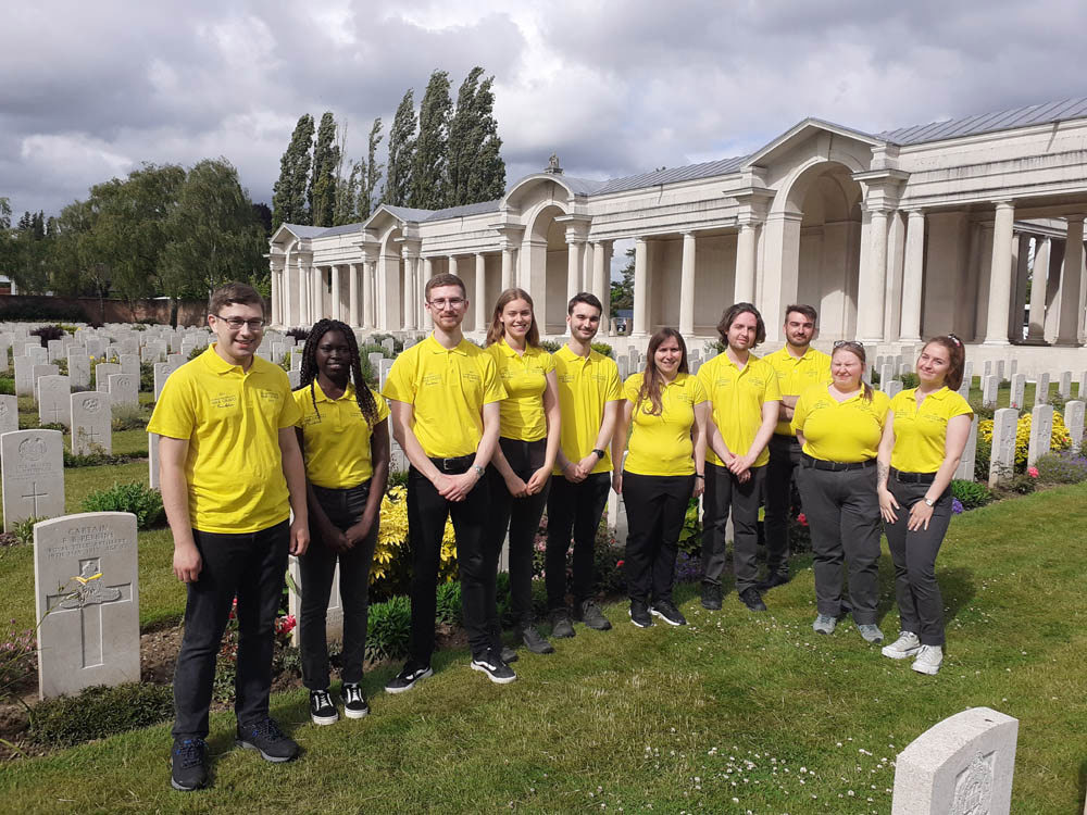 Group of CWGF Guides in bright yellow polo shirts pose in front of headstones in Faubourg D'Amiens War Cemetery, France.