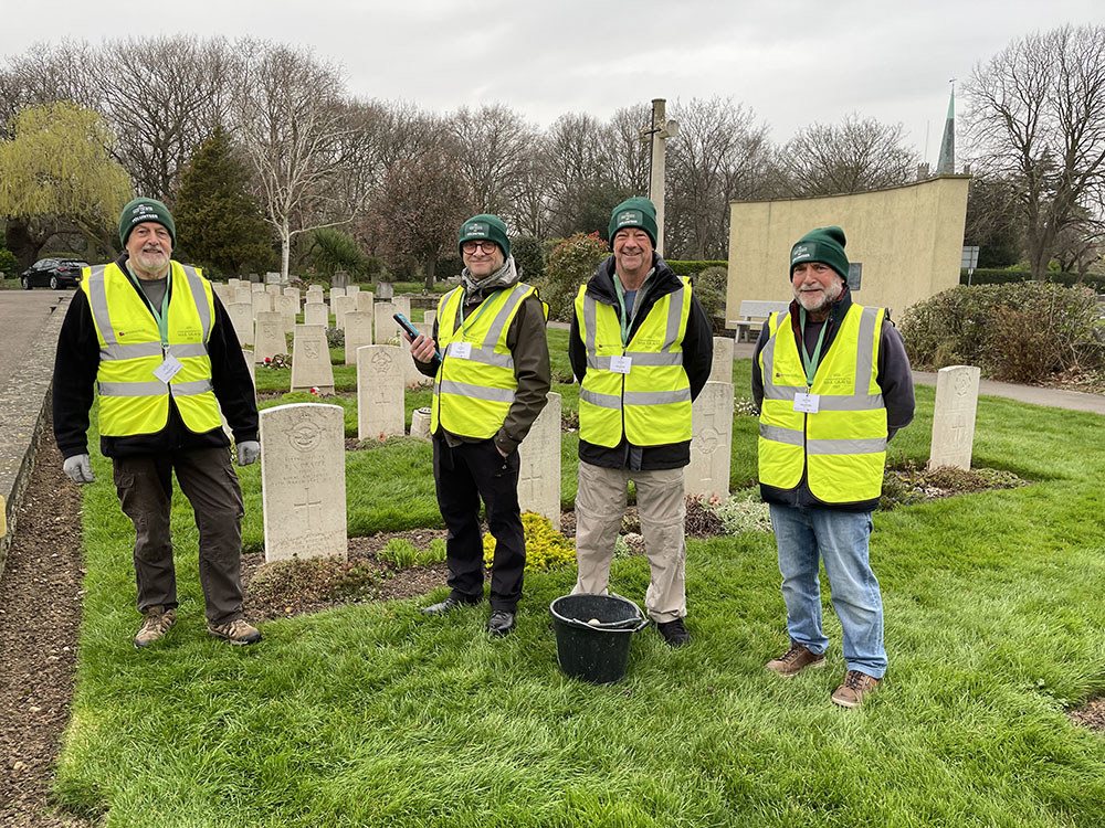 Four middle aged men in CWGC green woolly hats and hi-vis vets pose in front of war graves in a military cemetery.