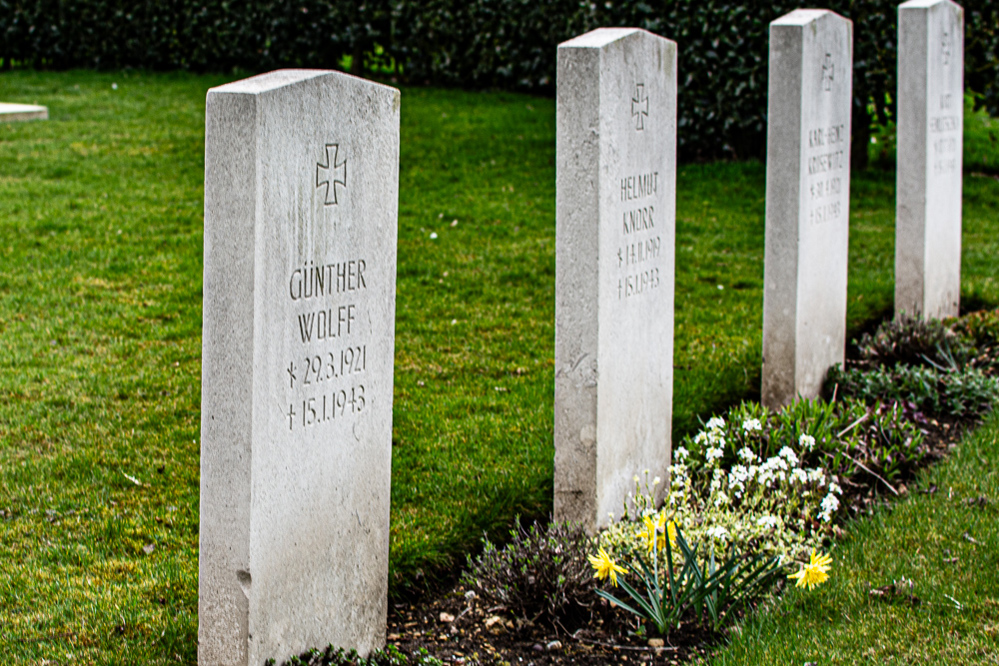 Row of German war graves at Scopwick burial ground. Each has a teutonic cross carved into the grey stone. Each headstone has a triangular pointed top. Daffodils and white flowers among other green plants are visible in the grave border.