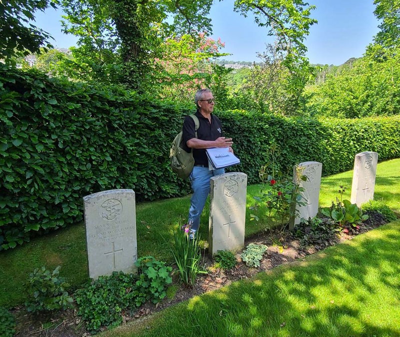 Tour guide speaking behind a row of CWGC headstones in a lush green cemetery on a summer's day