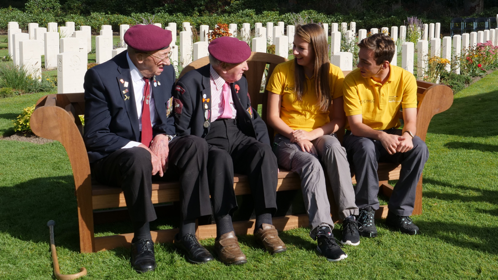 Two WW2 veterans speak with a young man and young woman on a bench in a CWGC cemetery.