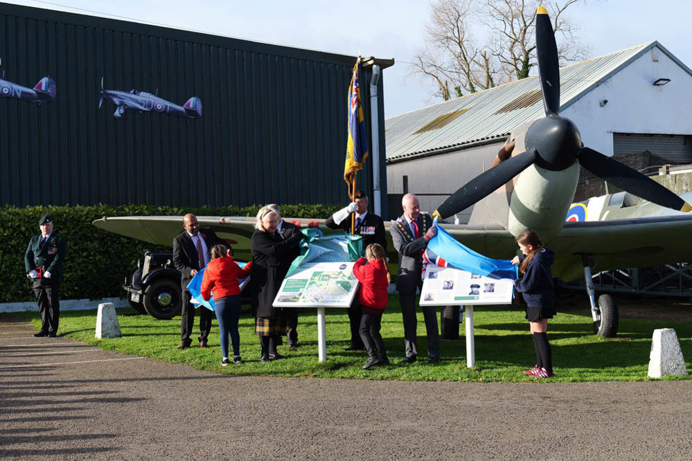 School children and dignitaries unveiling information panels in front of a replica Spitfire aircraft.