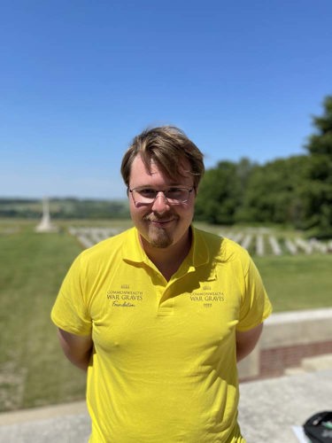 Roman poses in a yellow CWGF Guides polo shirt. CWGC  headstones and cross of sacrifice are visible in the background.