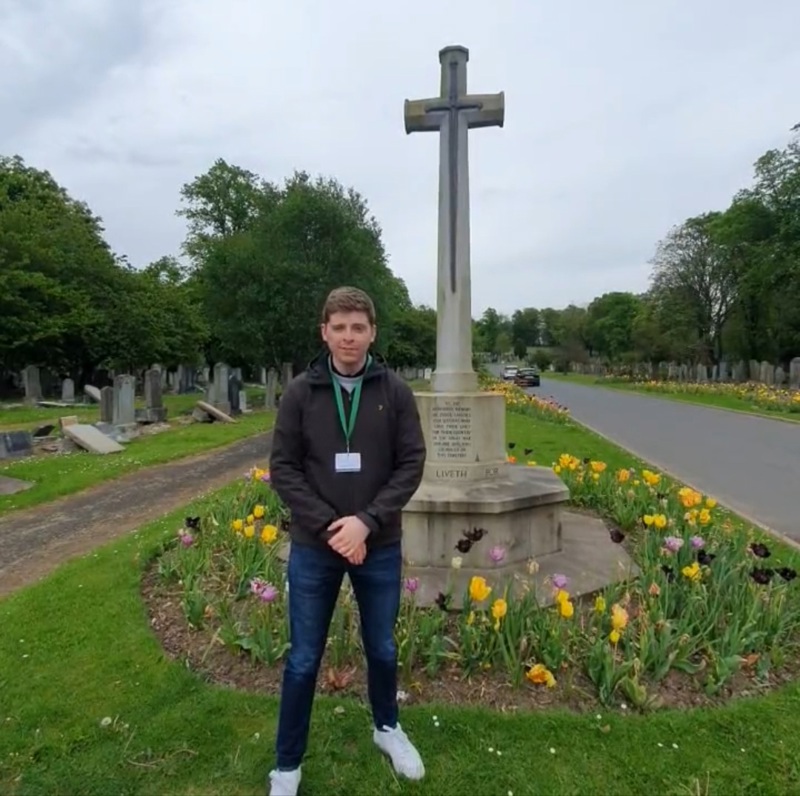 A young man poses in front of the Cross of Sacrifice within a Commonwealth War Graves cemetery.