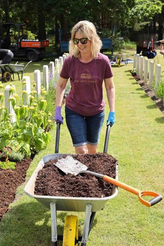 A middle aged blonde woman in sunglasses, maroon t shirt and jean shorts pushes a wheelbarrow full of much passed a row of CWGC headstones.