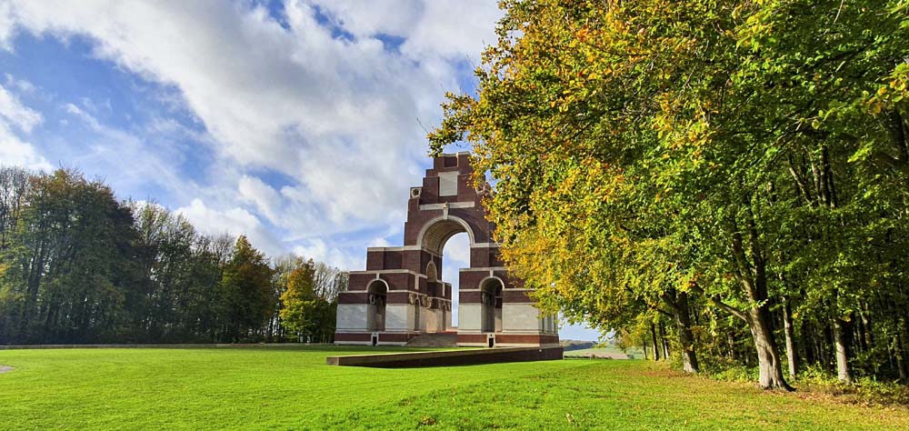 Thiepval Memorial sitting atop a vivid green lawn, flanked by tress showing a late summer golden-green hue. The sky is blue featuring thick white clouds.