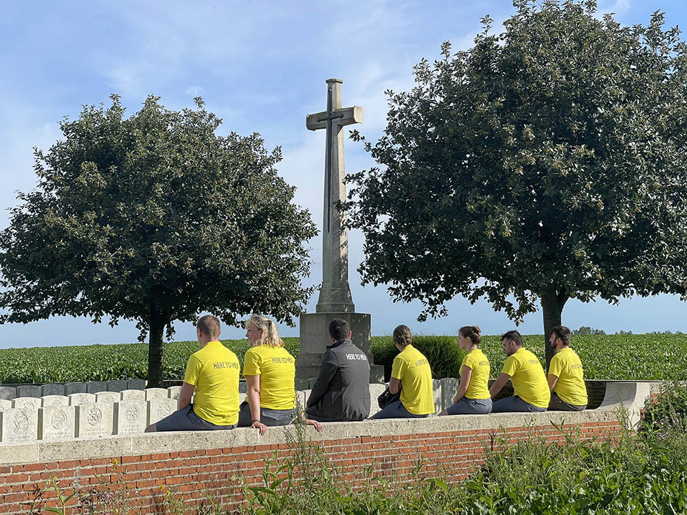 CWGF guides sitting on a wall of a CWGC cemetery. The wall is red brick. A cross of sacrifice, flanked by two trees, is visible in the background.