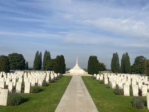 View of the Cross of Sacrifice at Tyne Cot Cemetery. A stone path flanked by green lawn and rows of headstones leads up to the Cross which is perched atop a white stone platfrom.