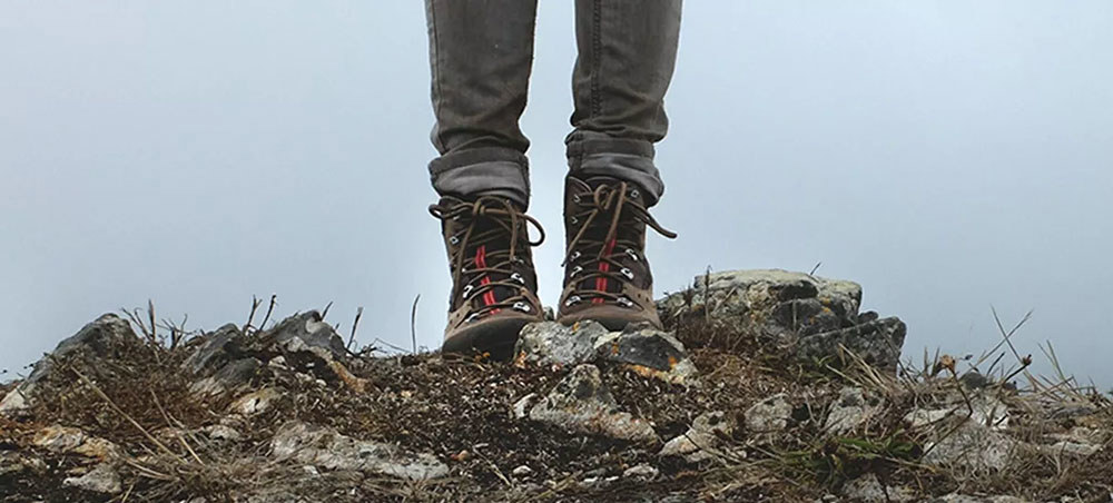 A shot of a walker's legs and feet showing his hiking boots. The figure is standing on a rocky outcrop against a blue-grey sky.