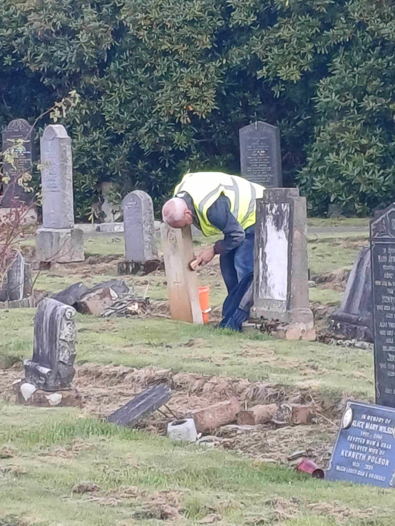 Man in a hi-vis vest scrubbing a CWGC headstone with a wire brush to clean it