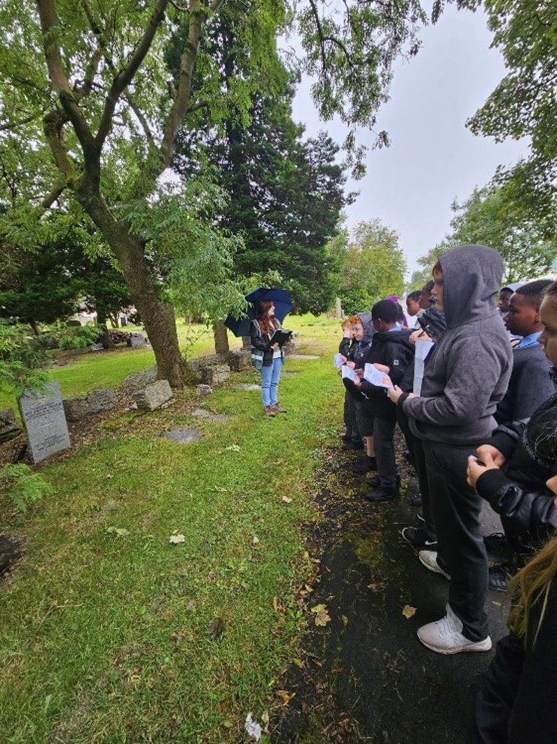 A young woman holding an umbrella gives a talk to a group of tour guides in a cemetery.