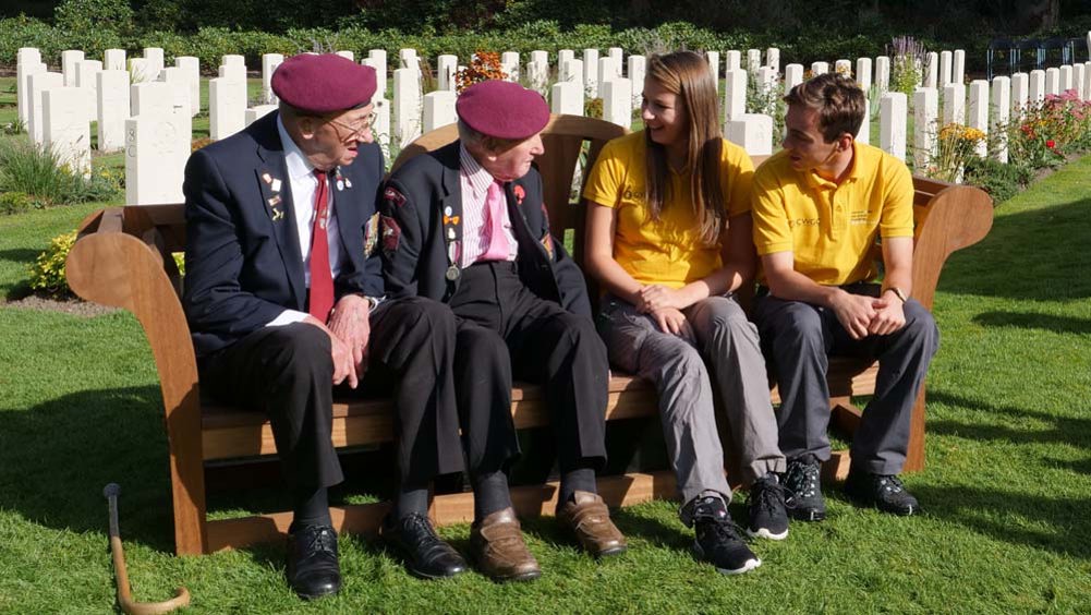 Two World War Two veterans in blazers, ties, and claret berets chat with two CWGF guides on a wooden bench. Rows of CWGF headstones can be seen in the background.