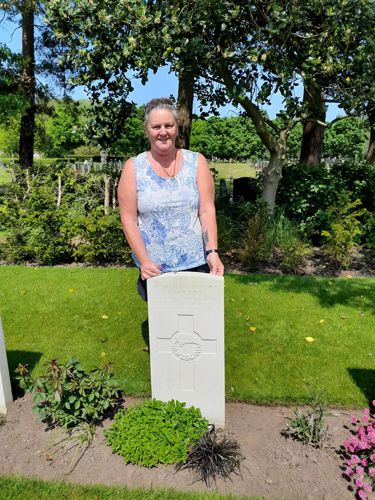 A middle aged woman with tied back grey hair, wearing a blue floral top, poses next to the CWGC headstone of Flight Sergeant Sidney Cains.