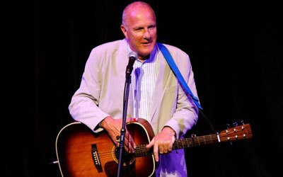 Richard Digense performs on stage. He is hlding a brown sunburst acoustic guitar with a black strap. Richard is standing in front of a microphone held on a stand.