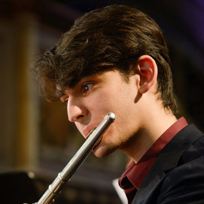 Daniel Swarni playing a silver metallic flute. He is wearing a black suit with a red shirt.