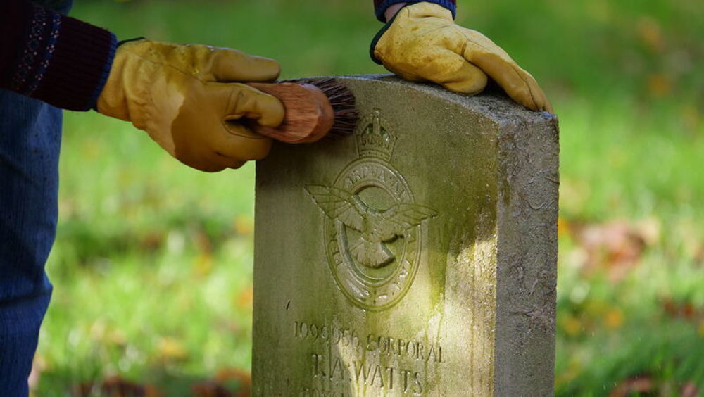 A close up of a man's hands cleaning a CWGC RAF headstone with a wire brush and some warm water.