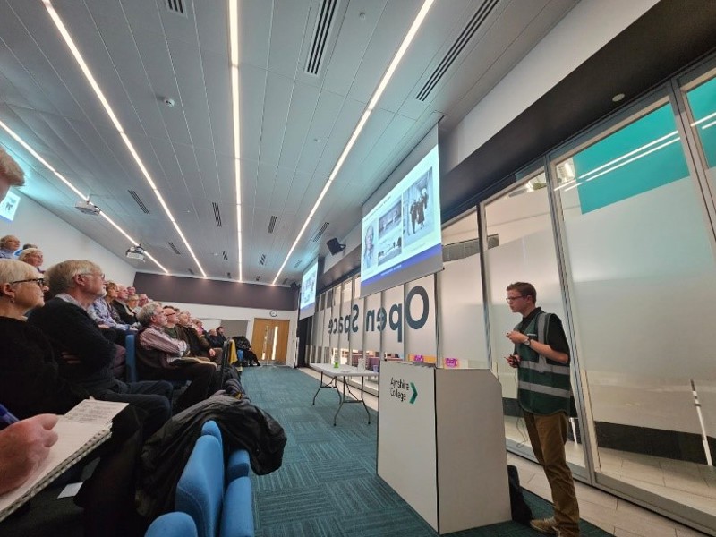 A young man in a green hi vis vest delivers a lecturer to an interested audience.