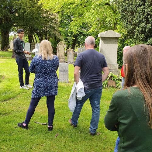 A Commonwealth War Graves volunteer speaker gives a tour of Greenock Cemetery. Around four visitors of various ages are listening attently to the speaker. Headstones can be seen in the background.