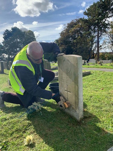 A middle-aged man wearing sunglasses and a hi-vis vest scrubs a CWGF headstone with a wire brush and some water to clean it.