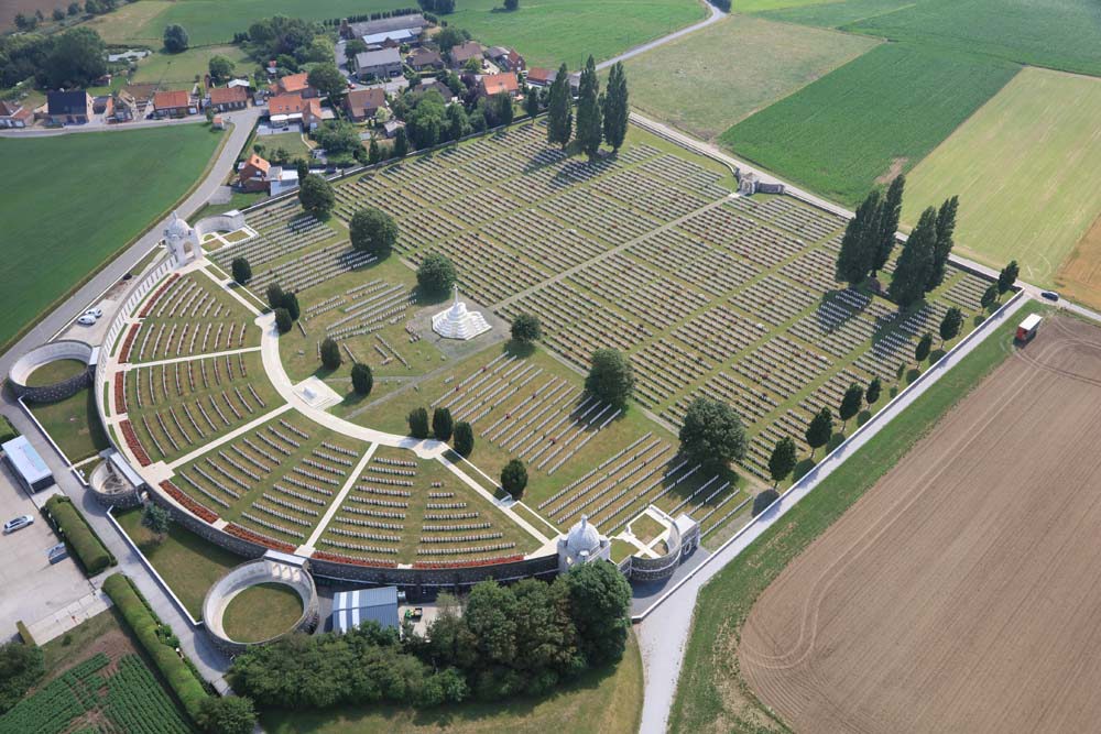 Overhead shot of Tyne Cot Cemetery displaying neatlyarrayed headstones, the curving wall of the Tyne Cot Memorial, and neatly trimmed landscaping.