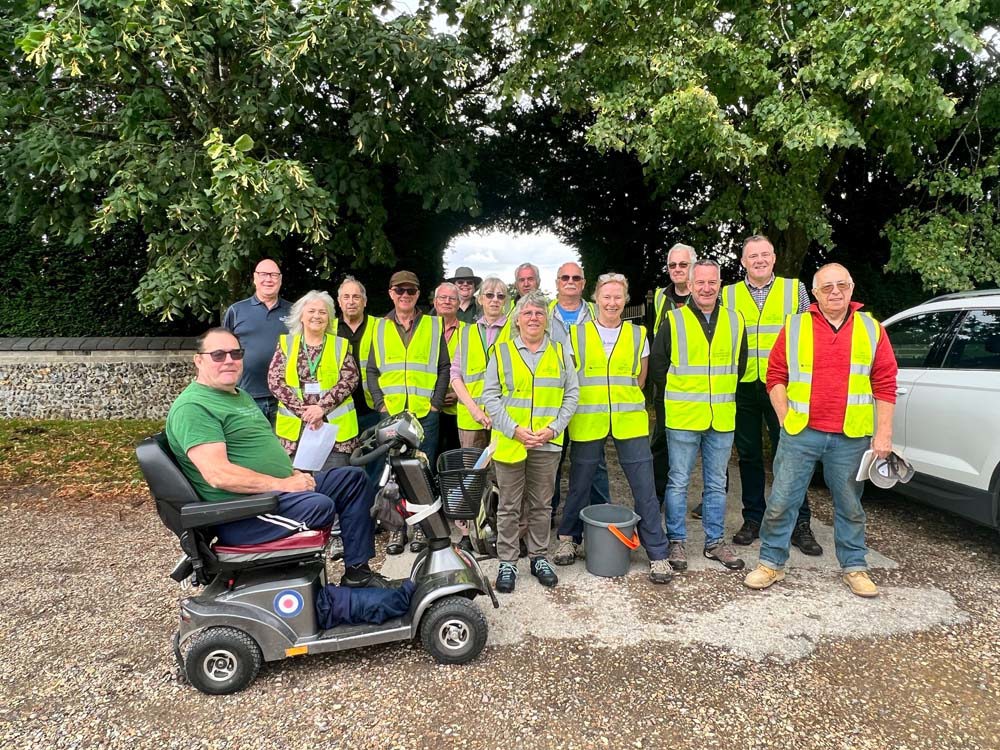 A group of middle aged people posing in hi-vis shirts in a carp park in front of a CWGC cemetery.