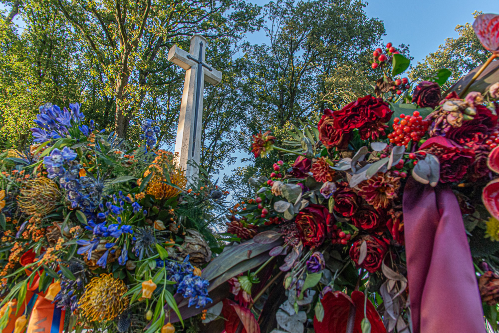 Bouquets of colourful flowers, featruing lilalcs, roses, magnolias and more bright blooms. A CWGC Cross of Sacrifice is visible in the background.