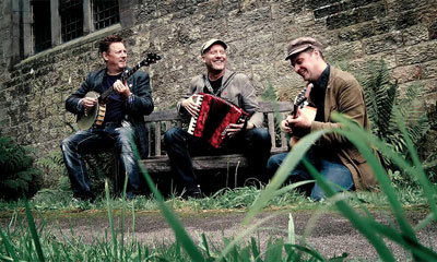 Folk trio Harp & a Monkey sit on a wooden bench in front of a church wall. Green blades of grass can be seen across the camera. From left to right, one musician is holding a banjor, the other an accordion, and the third is playing an acoustic guitar.