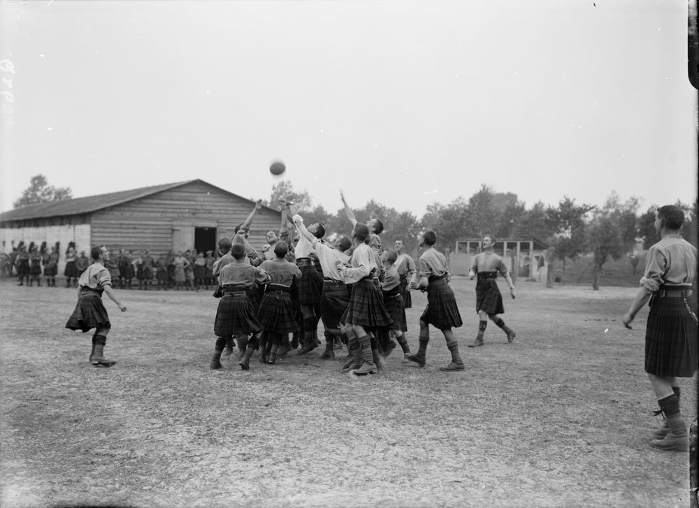 Black and white photo of Scots guardsmen from WW1 era playing a game of rugby. The players are in short sleeve shirts and wearing kilts. A wooden military hut can be seen in the background.