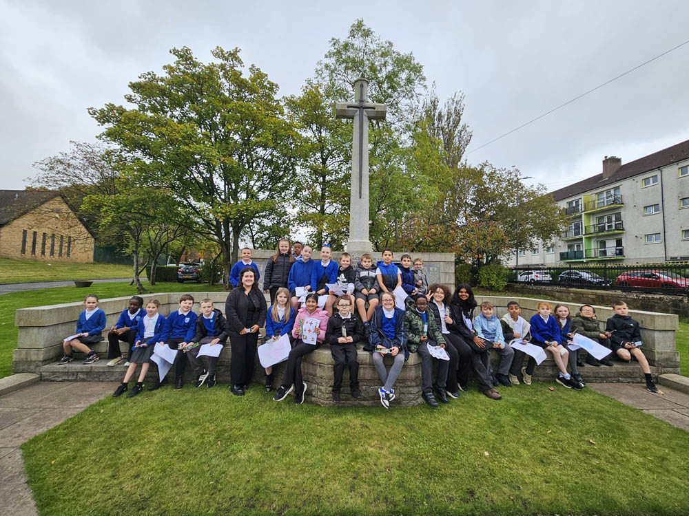 Primary school children and University of Glasgow students pose on the Cross of Sacrifice in a Scottish cemetery.