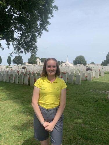 Portrait photo of Erin Harris in her yellow CWGF polo and grey shorts. She is standing in front of the graves at Tyne Cot Cemetery.