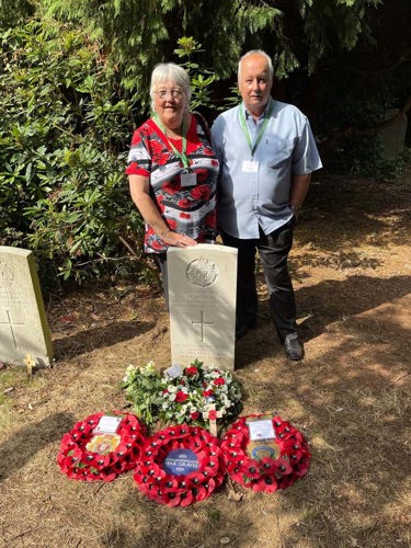 Jan & Len Pritchard pose next to a CWGF headstone Jan is wearing a bright poppy patterned top with Len in a blue short sleeve shirt. Several red poppy wreathes are laid at the base of the white limestone headstone.