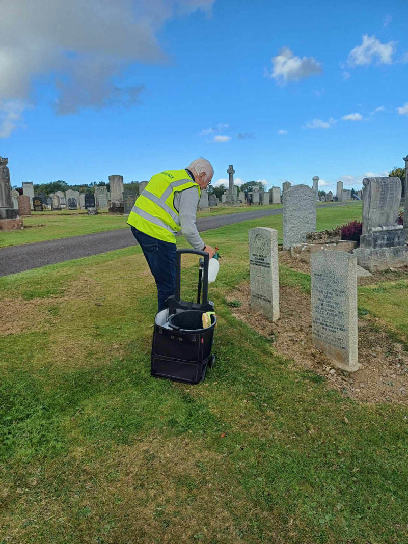 A man in a high vis vest sprays a CWGC headstone with water to clean it.