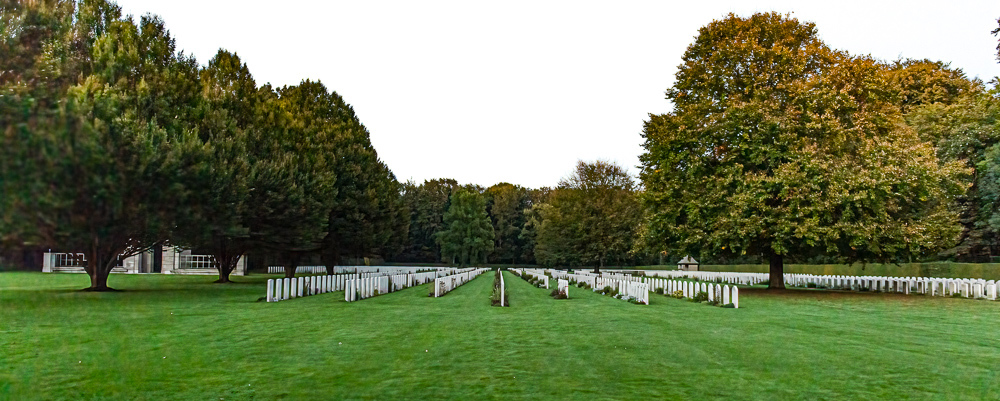 Reichswald War Cemetery at dusk with rows of CWGC headstones flanked by tall oak trees.