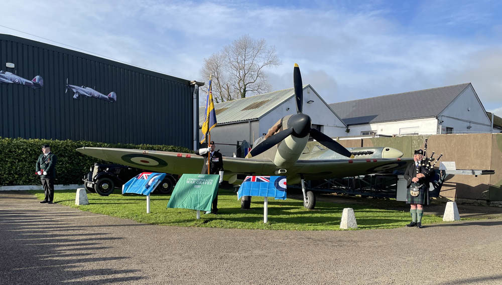 A piper stands in front a replica spitfire, playing the bagpipes. To his left stands a standard bearer. Another honour guard can be seen to the far left.